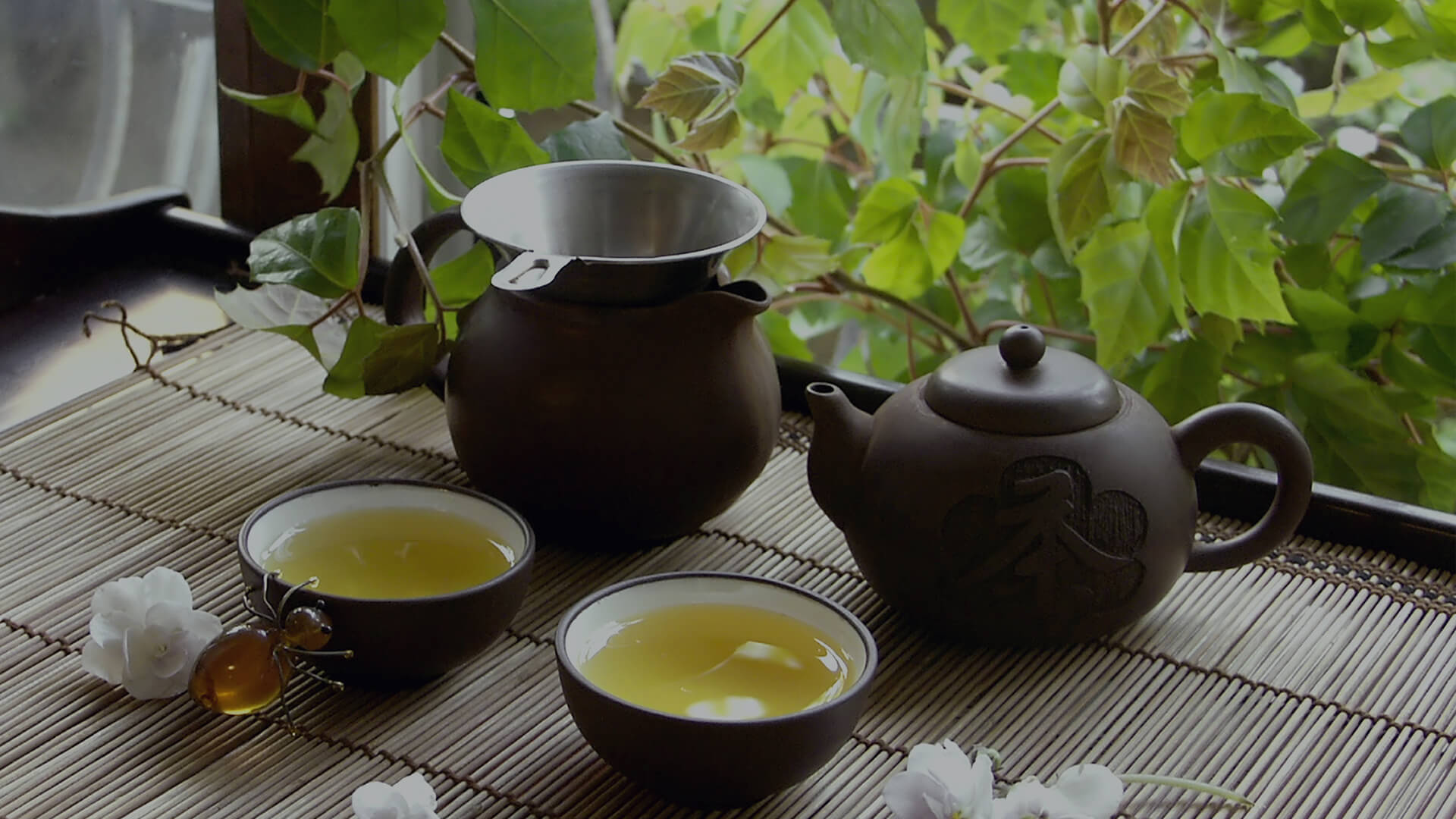 Zestaw do herbaty z dwoma czarkami i ciemnymi ceramicznymi czajnikami na bambusowej macie, w tle zielone liście przy oknie.