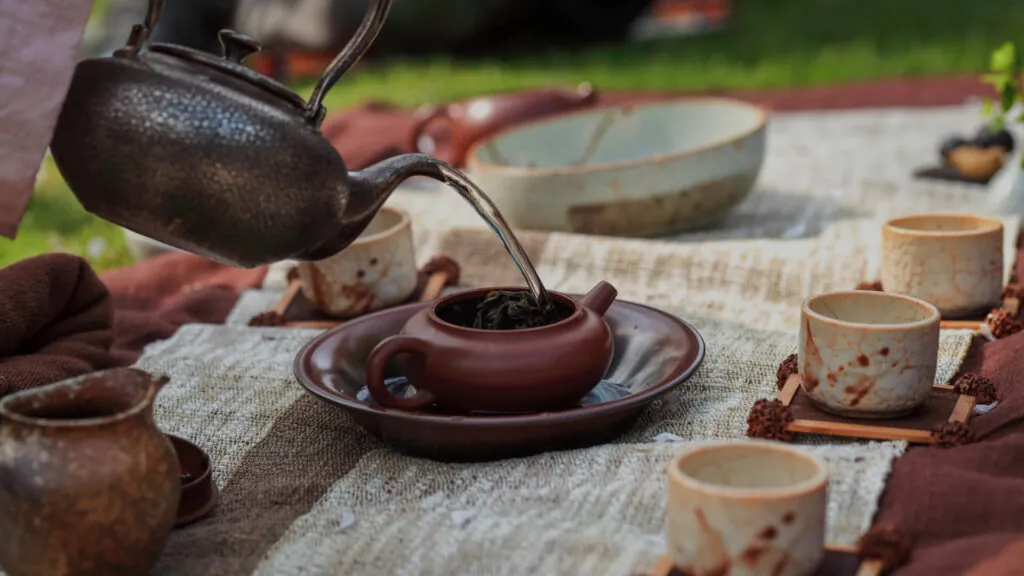 Koc rozłożony na trawie, na kocu tradycyjne filiżanki. Do jednej z filiżanek nalewana jest woda z czajnika.
