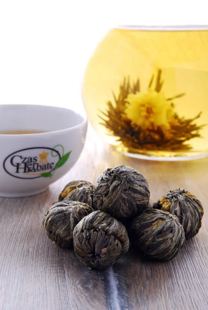 Filiżanka z napisem Czas na Herbatę, kulki z herbatą kwitnącą oraz szklany czajnik z zaparzoną herbatą kwitnącą.