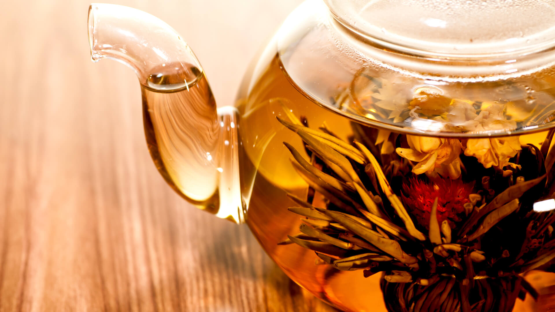 Szklany dzbanek z herbatą kwitnącą, zanurzoną w naparze, na jasnobrązowym drewnianym blacie.