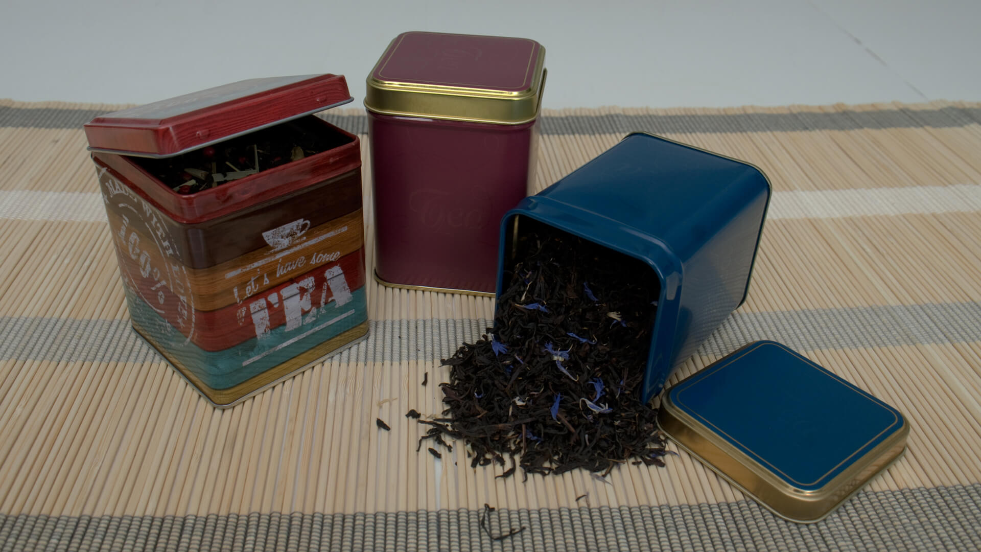 Kolorowe puszki na herbatę, jedna przewrócona z rozsypaną czarną herbatą z niebieskimi płatkami, ustawione na bambusowej macie.