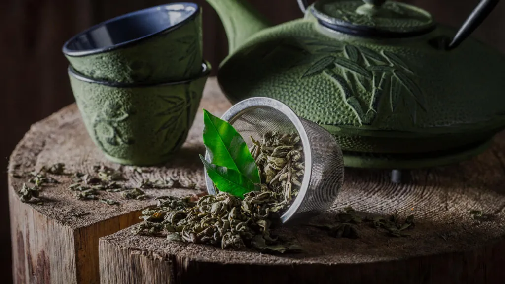 Zielony żeliwny czajnik z orientalnym wzorem, filiżanki, sitko z liśćmi zielonej herbaty i świeżymi listkami, wszystko ustawione na drewnianym pieńku.