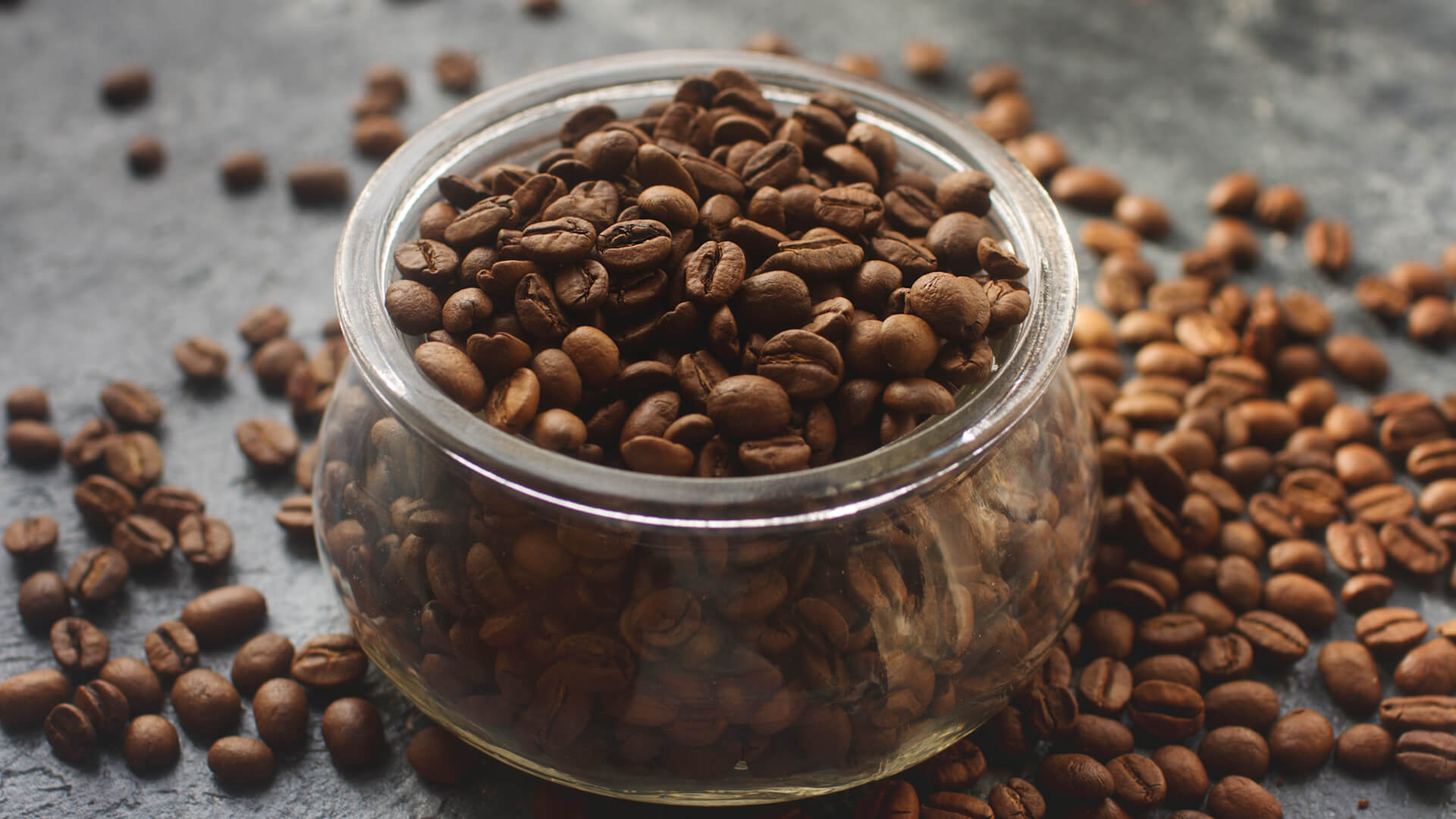 Szklany słoik pełny ziaren kawy ustawiony na ciemnym blacie, wokół rozsypane ziarna.