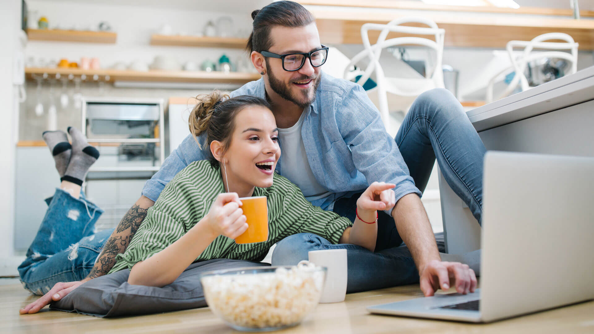 Para relaksująca się w domowej kuchni, leżąca na podłodze z laptopem, kubkami i miską popcornu podczas wspólnego oglądania lub pracy.