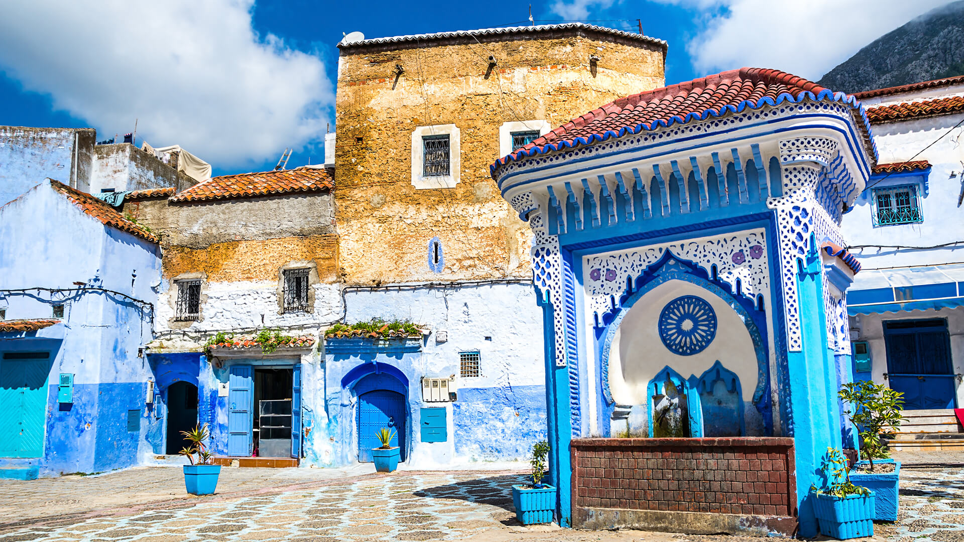 Budynek z fontanną zdobioną błękitną mozaiką w Maroko, otoczone niebieskimi ścianami, jasnymi donicami i żółtą fasadą pod błękitnym niebem.