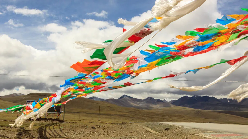 Kolorowe tybetańskie flagi modlitewne powiewające na wietrze, rozciągnięte nad górskim pustkowiem pod zachmurzonym niebem.