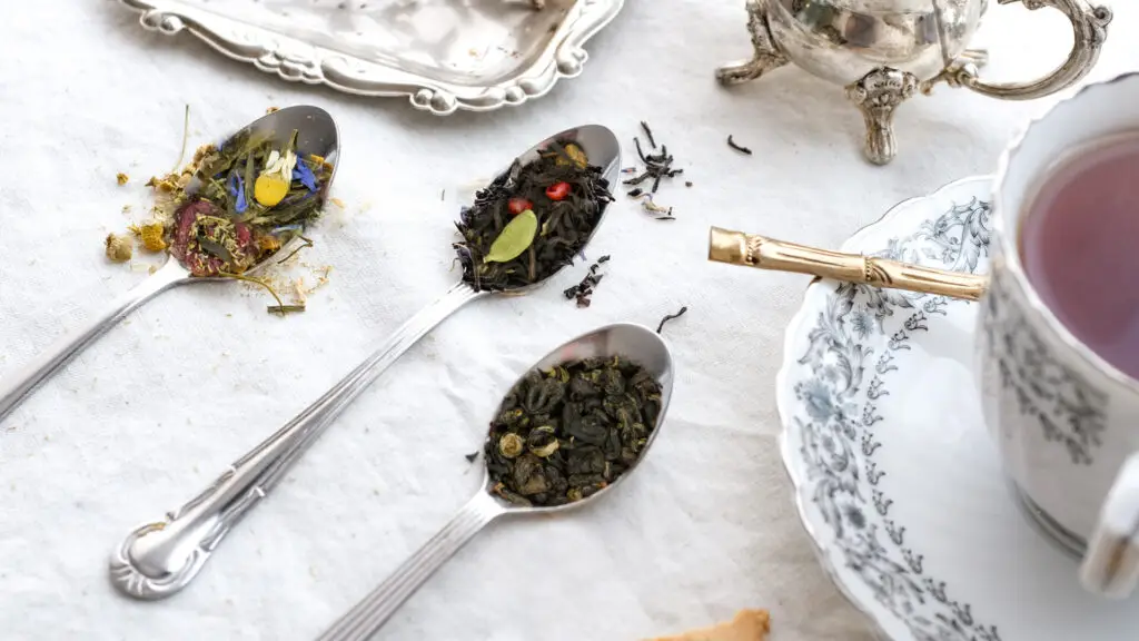 Trzy łyżeczki z różnymi rodzajami herbaty liściastej na białym obrusie, obok filiżanka z herbatą w porcelanie.