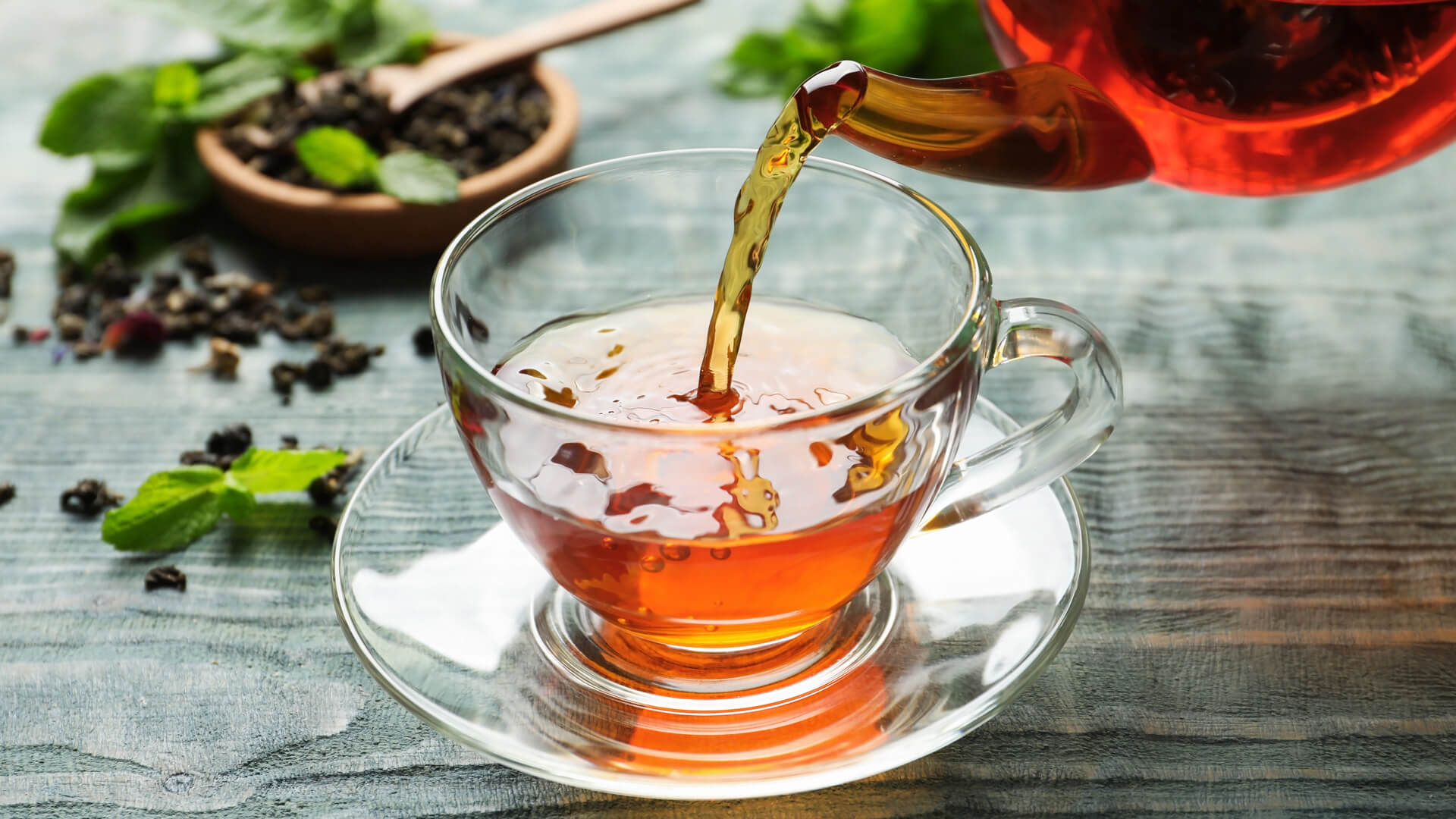 Nalewanie świeżo zaparzonej czarnej herbaty do szklanej filiżanki na spodku, obok listki mięty i sypana herbata na drewnianym tle.