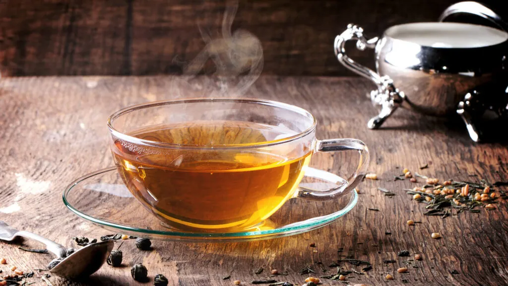 Szklana filiżanka z gorącą herbatą ustawiona na spodku, wokół rozsypane liście herbaty i zioła, w tle srebrne naczynie na drewnianym blacie.