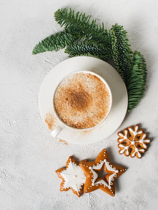 Biała filiżanka z kawą, gałązka drzewa iglastego i udekorowane pierniki.