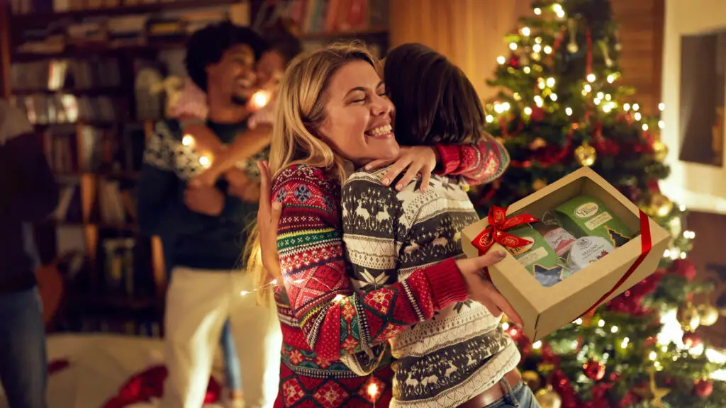 Przytulające się osoby w świątecznych swetrach, jedna trzyma prezentowy zestaw herbaty przy choince, domowa atmosfera.