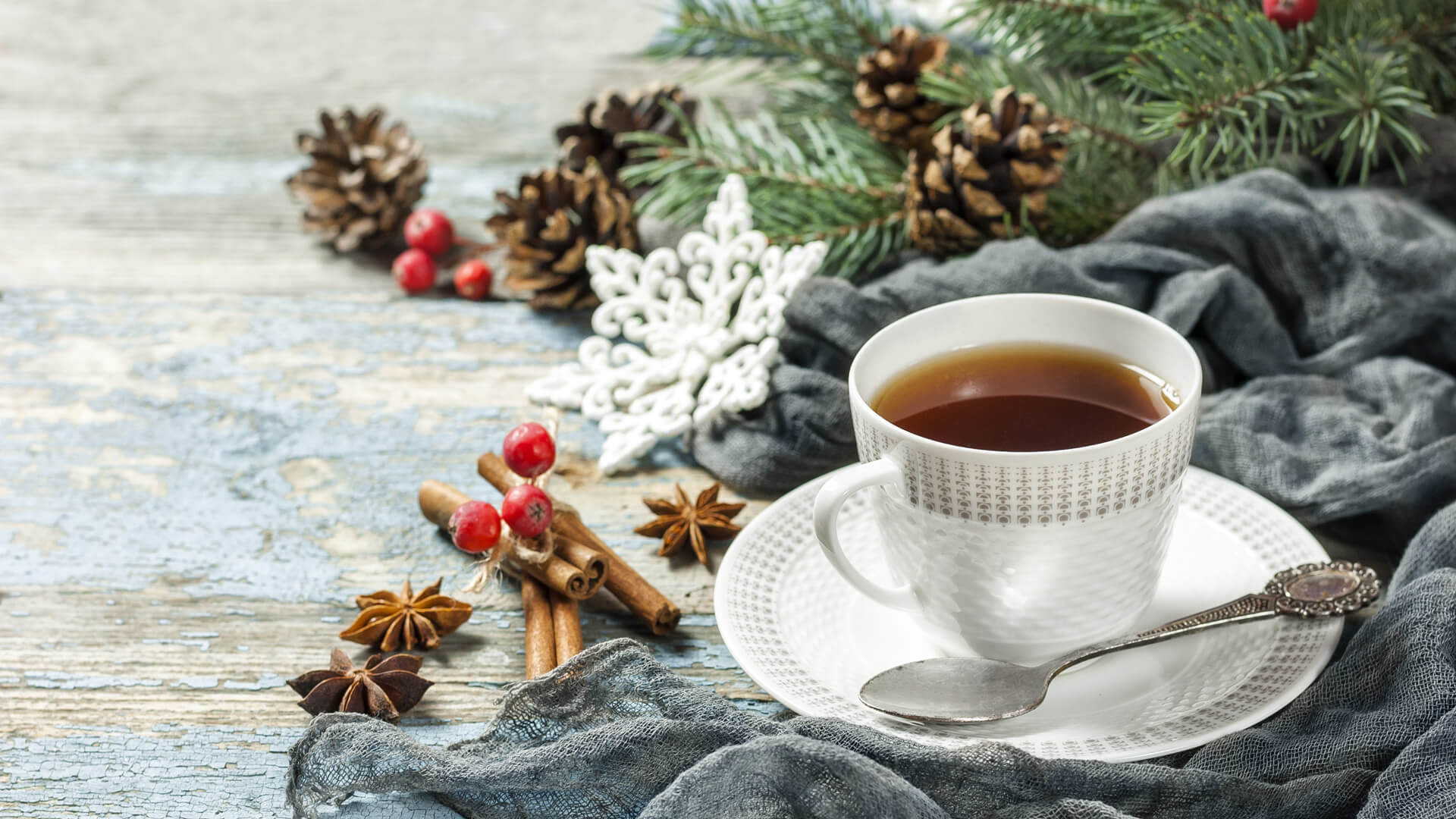 Biała filiżanka z herbatą na spodku, obok gałązki igliwia, szyszki, anyż, laski cynamonu, owoce i ozdobna śnieżynka na błękitnym drewnianym tle.