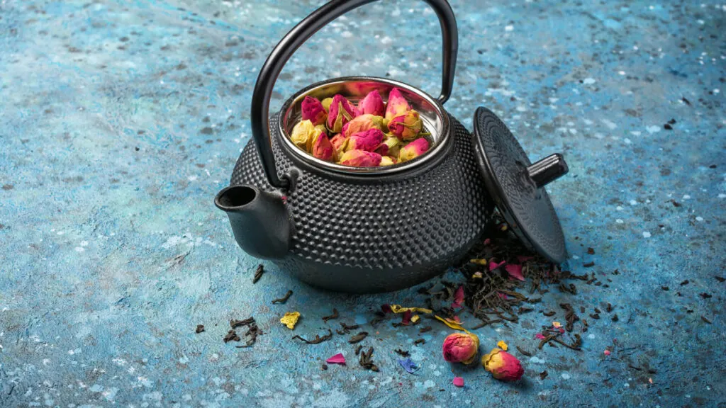 Czarny żeliwny czajnik z otwartą pokrywką, wypełniony różanymi pąkami i sypaną herbatą, na niebieskim tle, wokół rozsypane płatki oraz kawałki suszu.