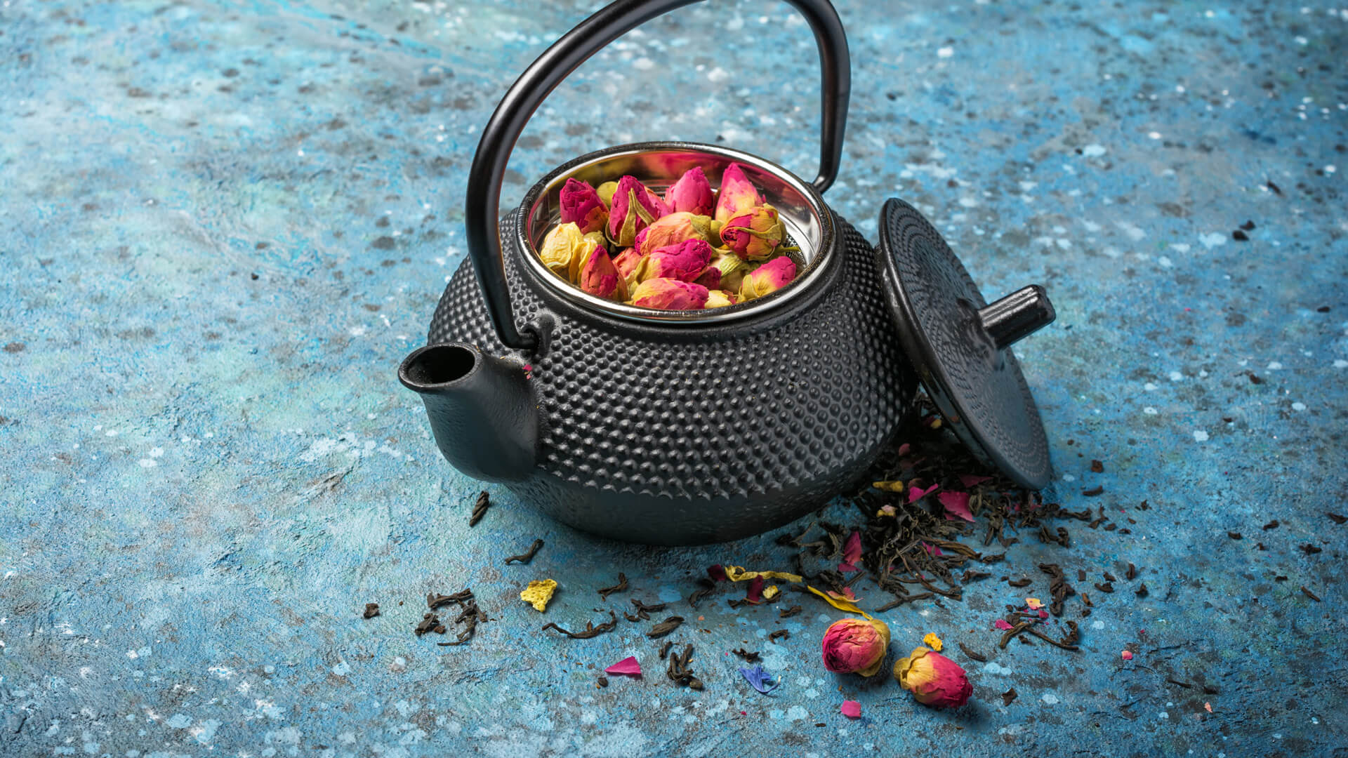 Czarny żeliwny czajnik z otwartą pokrywką, wypełniony różanymi pąkami i sypaną herbatą, na niebieskim tle, wokół rozsypane płatki oraz kawałki suszu.