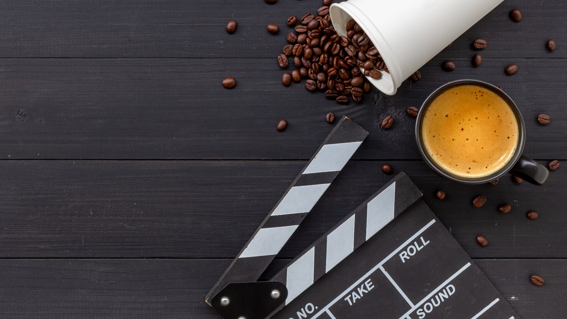 Czarna filiżanka z kawą, rozsypane ziarna i przewrócony biały kubek jednorazowy, obok czarno-biała klaps filmowy na ciemnym drewnianym tle.