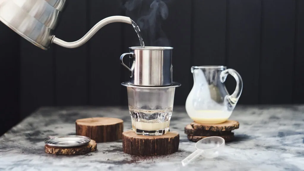 Gorąca woda nalewana z czajnika do metalowego zaparzacza na szklance z mlekiem skondensowanym, obok dzbanek z mlekiem i drewniane podstawki.