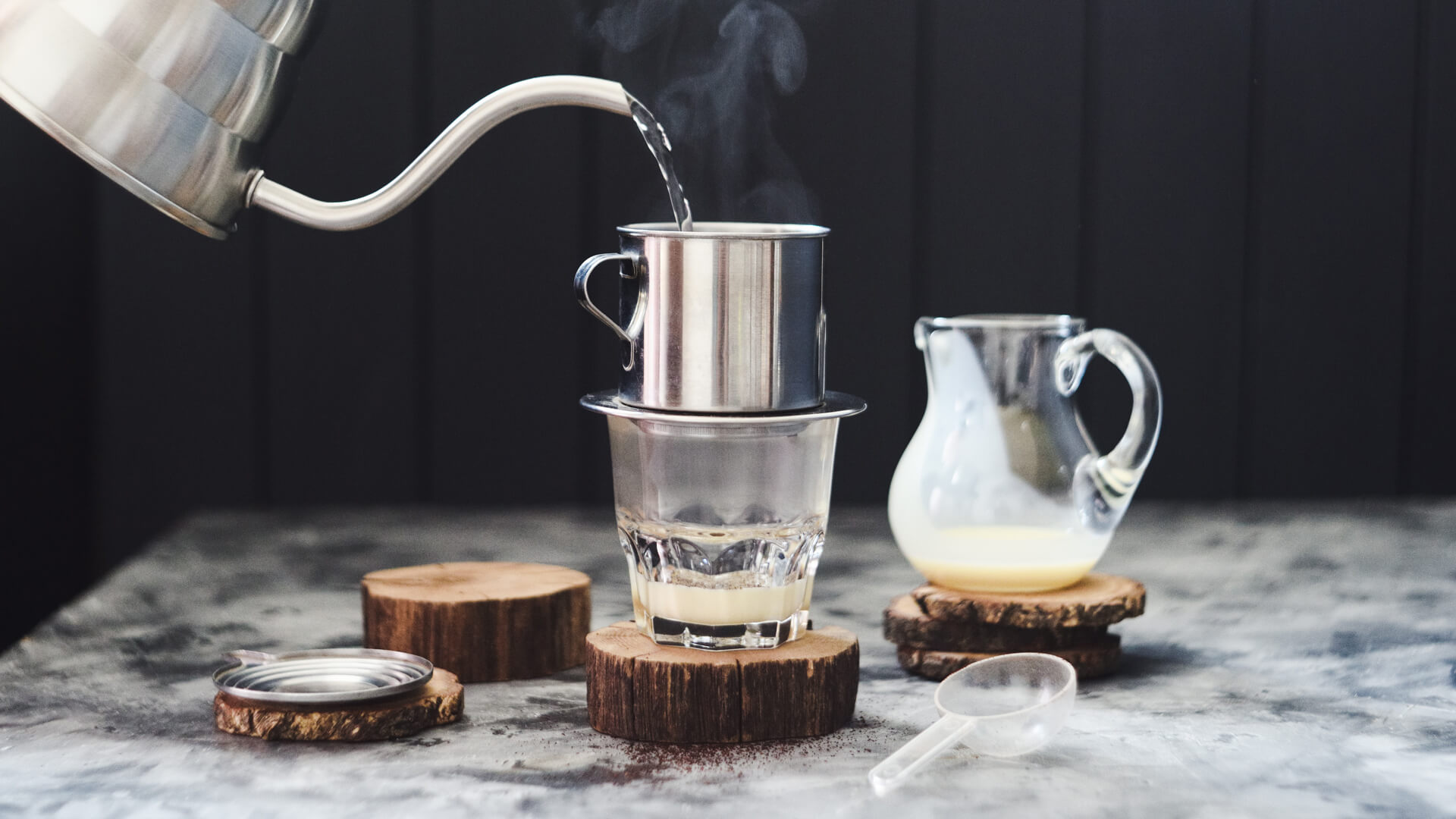 Gorąca woda nalewana z czajnika do metalowego zaparzacza na szklance z mlekiem skondensowanym, obok dzbanek z mlekiem i drewniane podstawki.