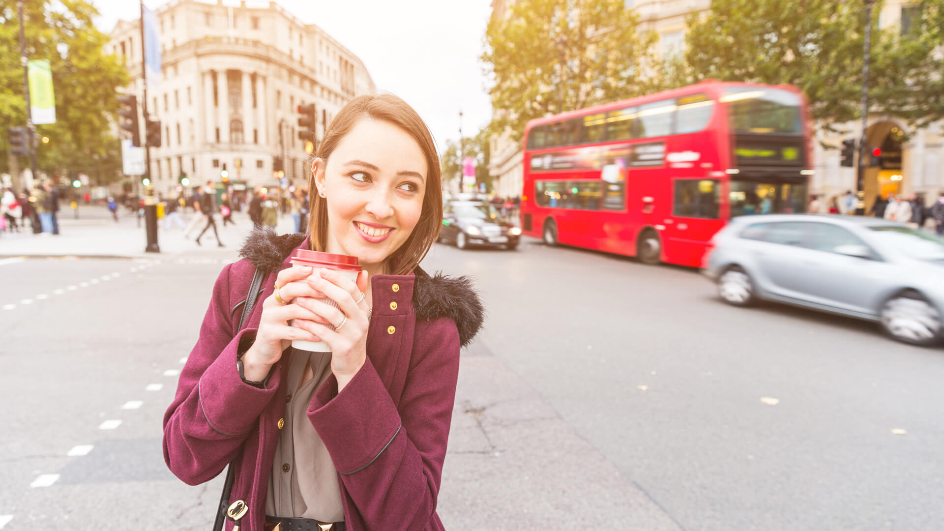 Osoba w bordowym płaszczu trzymająca kubek z napojem, stojąca na ulicy w Londynie z czerwonym piętrowym autobusem w tle i ruchem miejskim.