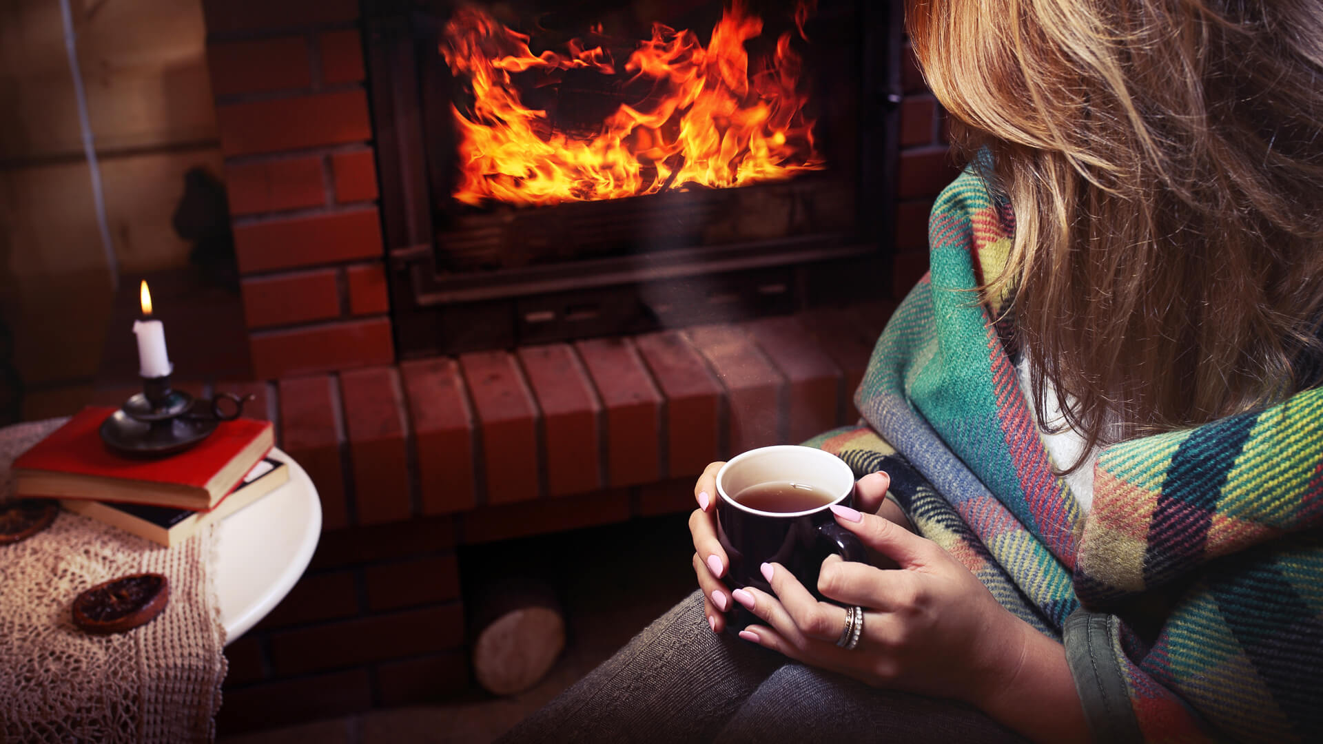 Osoba siedząca pod kocem z kubkiem gorącego napoju, przytulna atmosfera i płonący kominek, na stoliczku książki, świeca i ciasteczka.
