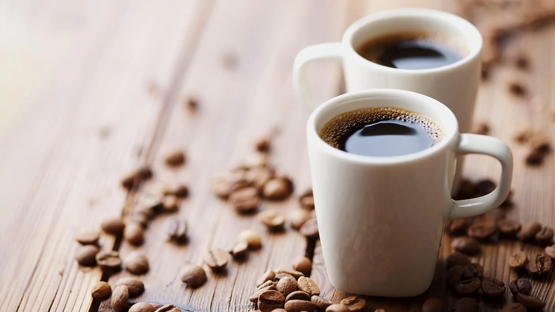Dwie białe kubki z czarną kawą ustawione na drewnianym stole, wokół rozsypane ziarna kawy.