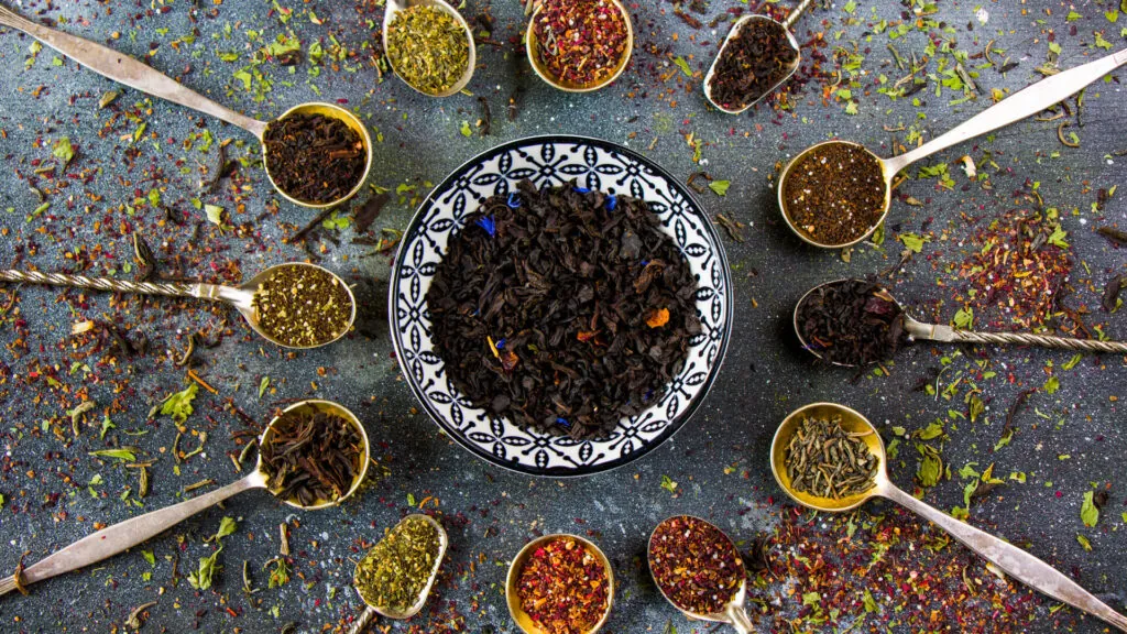 Różne rodzaje sypanej herbaty w łyżeczkach rozmieszczonych wokół miseczki z czarną herbatą, na dekoracyjnej powierzchni.
