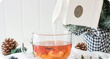 Nalewanie mleka z dzbanka do szklanej filiżanki z herbatą na tle zimowej dekoracji z szyszkami i gałązkami