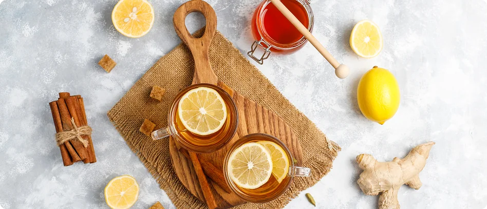 Widok z góry na dwie szklanki herbaty z plasterkami cytryny na drewnianej desce, wokół cynamon, miód, imbir i cytryna.
