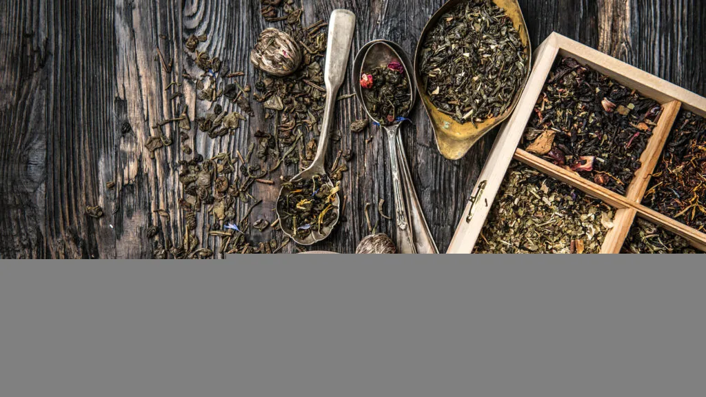 Różne rodzaje sypanej herbaty w drewnianej skrzynce i metalowych łyżkach na drewnianym stole, obok porcje suszu i miseczka.