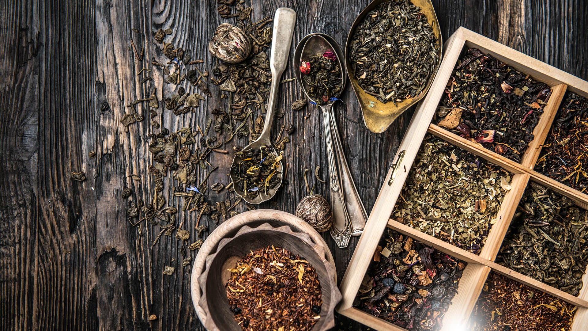 Różne rodzaje sypanej herbaty w drewnianej skrzynce i metalowych łyżkach na drewnianym stole, obok porcje suszu i miseczka.