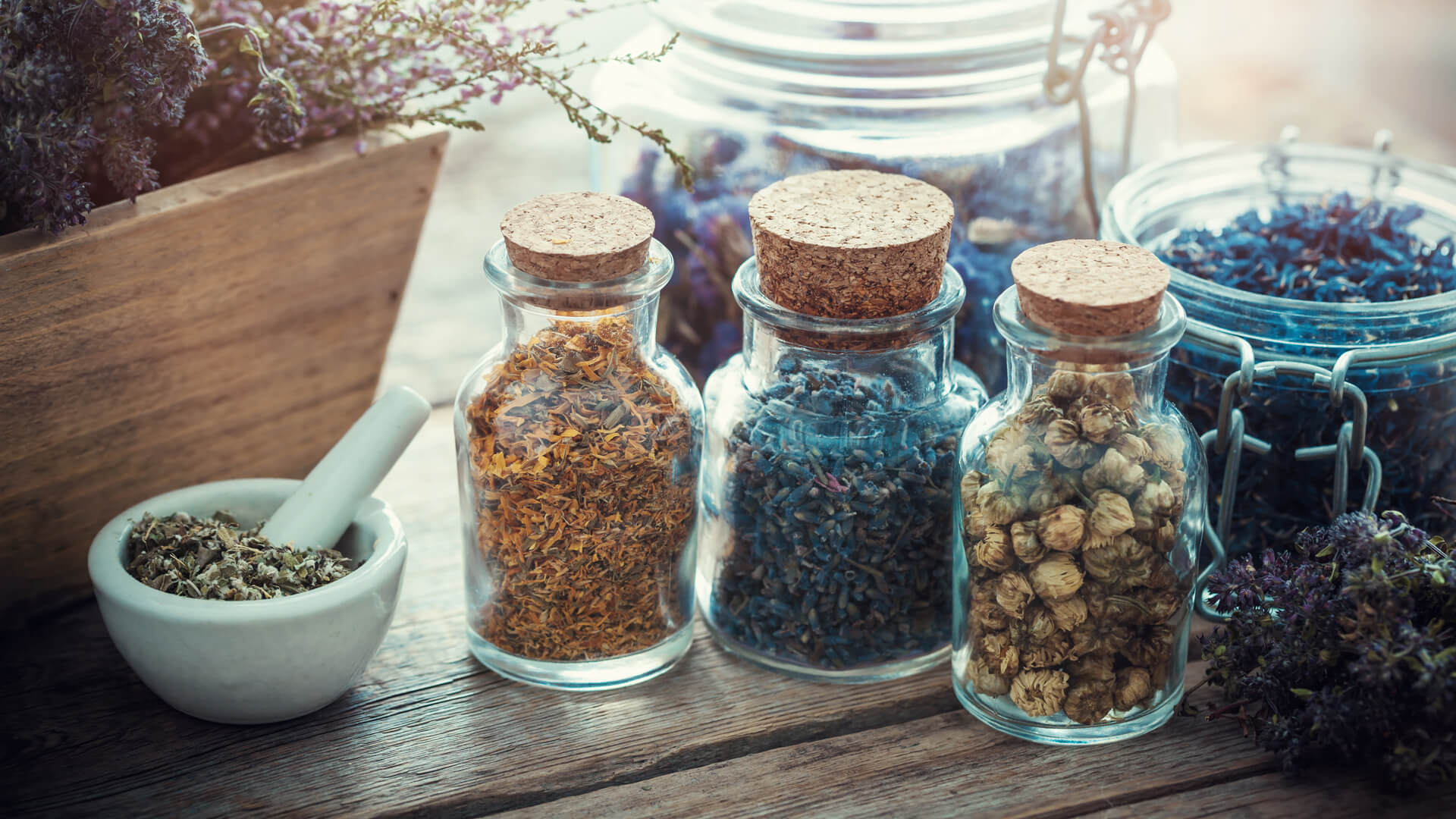 Szklane słoiki zamknięte korkami wypełnione suszonymi ziołami i kwiatamina drewnianym blacie; obok moździerz z mieszanką oraz bukiet lawendy.