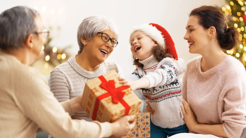 Rodzina wymieniająca się świątecznymi prezentami, dziecko w czapce Mikołaja sięga po zapakowane pudełko z czerwoną wstążką, w tle choinka i dekoracje.