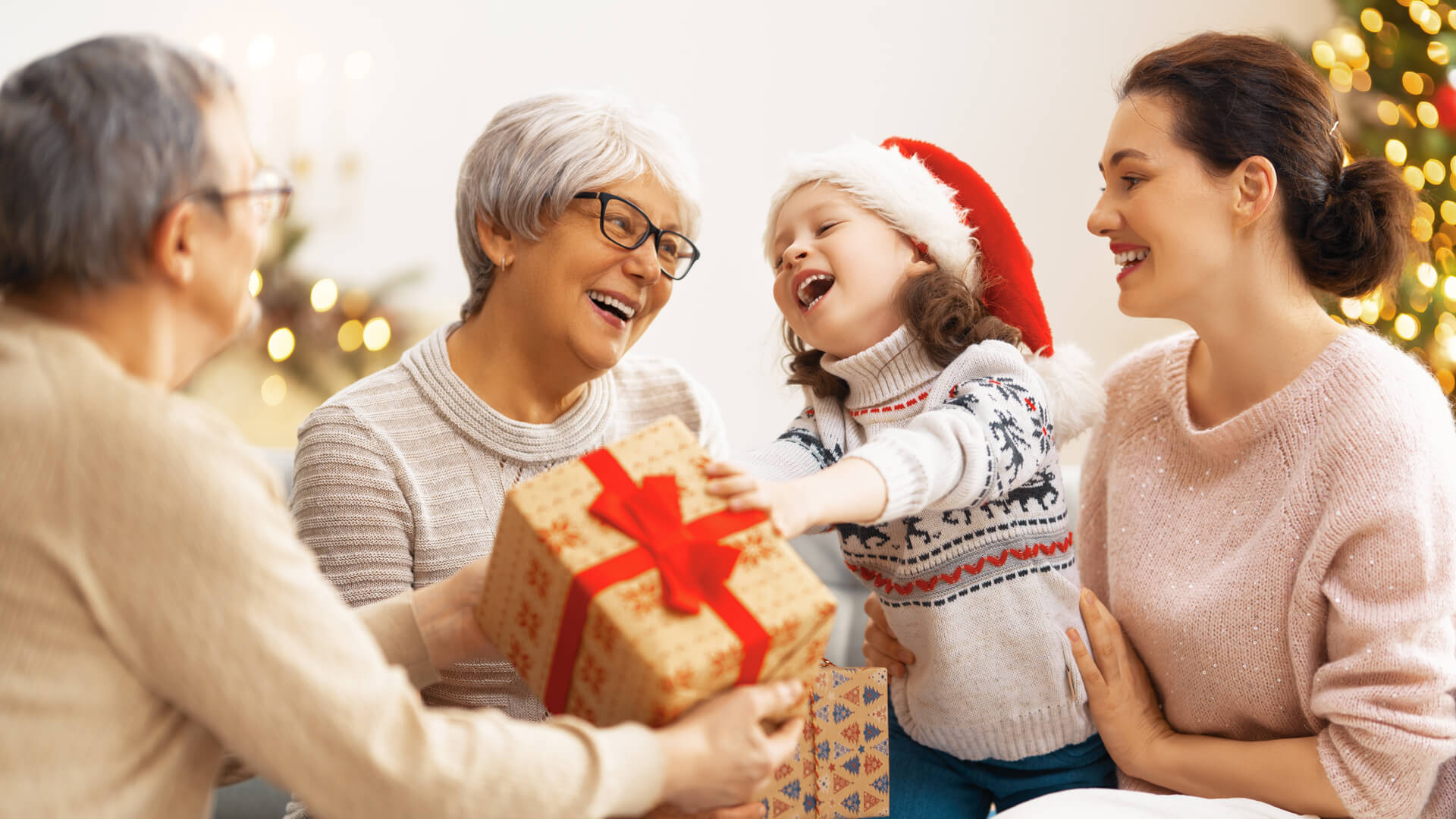 Rodzina wymieniająca się świątecznymi prezentami, dziecko w czapce Mikołaja sięga po zapakowane pudełko z czerwoną wstążką, w tle choinka i dekoracje.