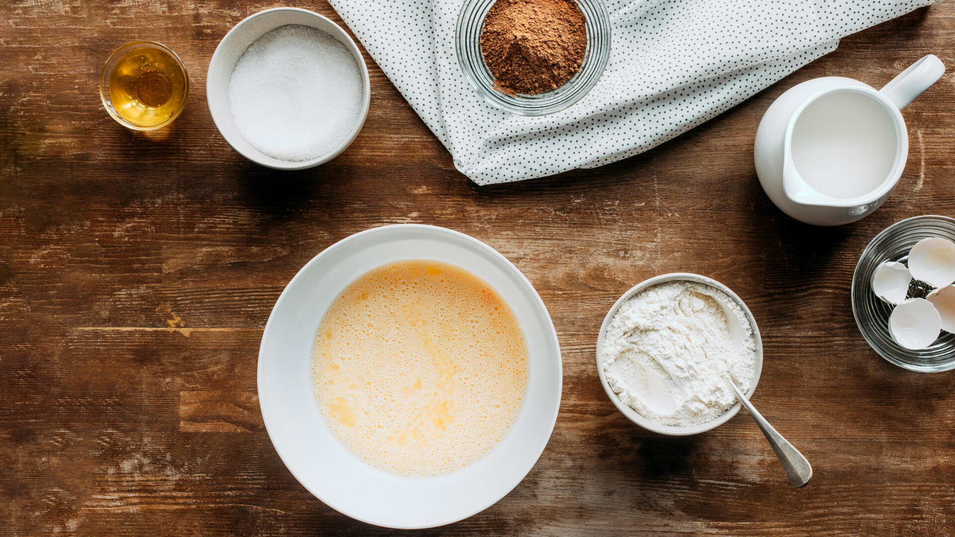 Składniki do wypieków rozmieszczone na drewnianym blacie: miska z roztrzepanymi jajkami, mąka, cukier, kakao, mleko, miód i skorupki jajek.