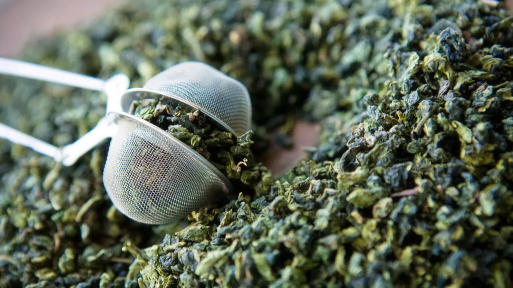 Zielona herbata liściasta rozsypana na stole, metalowe sitko zaparzacza wypełnione liśćmi.