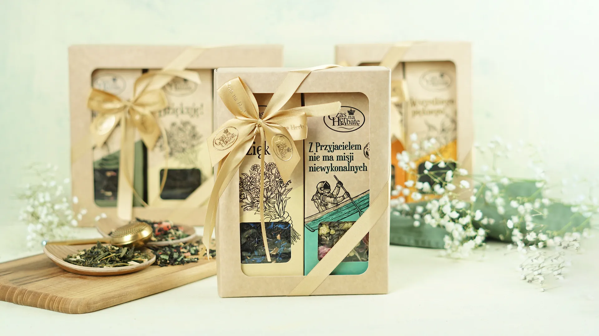 Zestaw prezentowy herbaty w ozdobnym pudełku z kokardą, w tle inne podobne opakowania i kwiaty.