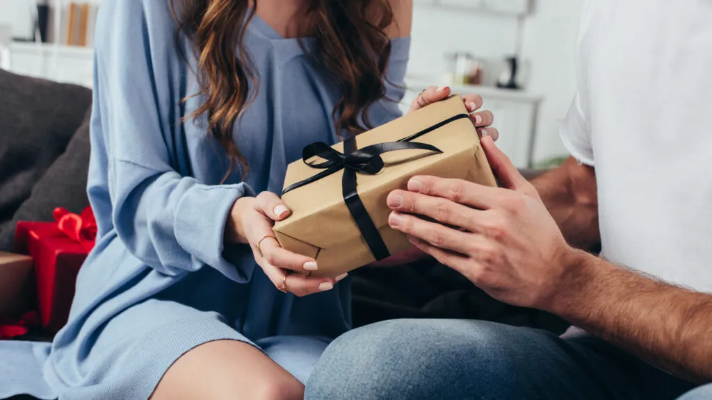 Dwie osoby przekazują sobie prezent zapakowany w brązowy papier z czarną wstążką.