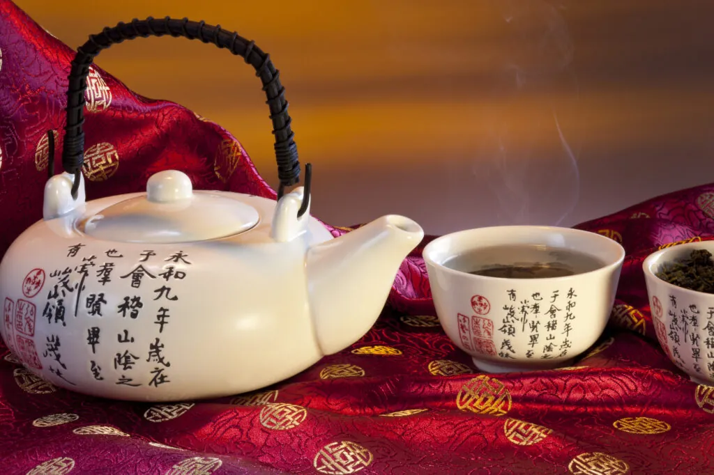 Biały czajnik i filiżanka z chińskimi znakami, parująca herbata ustawiona na czerwonym, zdobionym materiale.