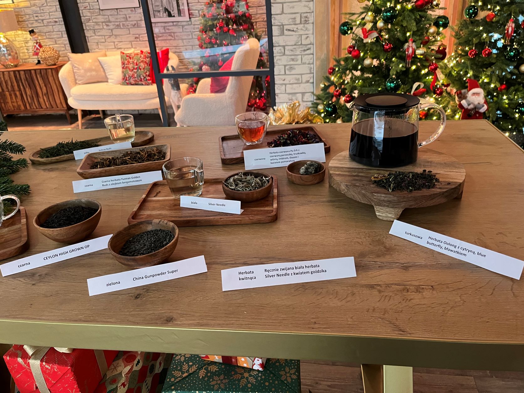 Stół z różnymi rodzajami herbaty i czajniczkiem, w tle świątecznie udekorowane studio telewizyjne z choinkami.