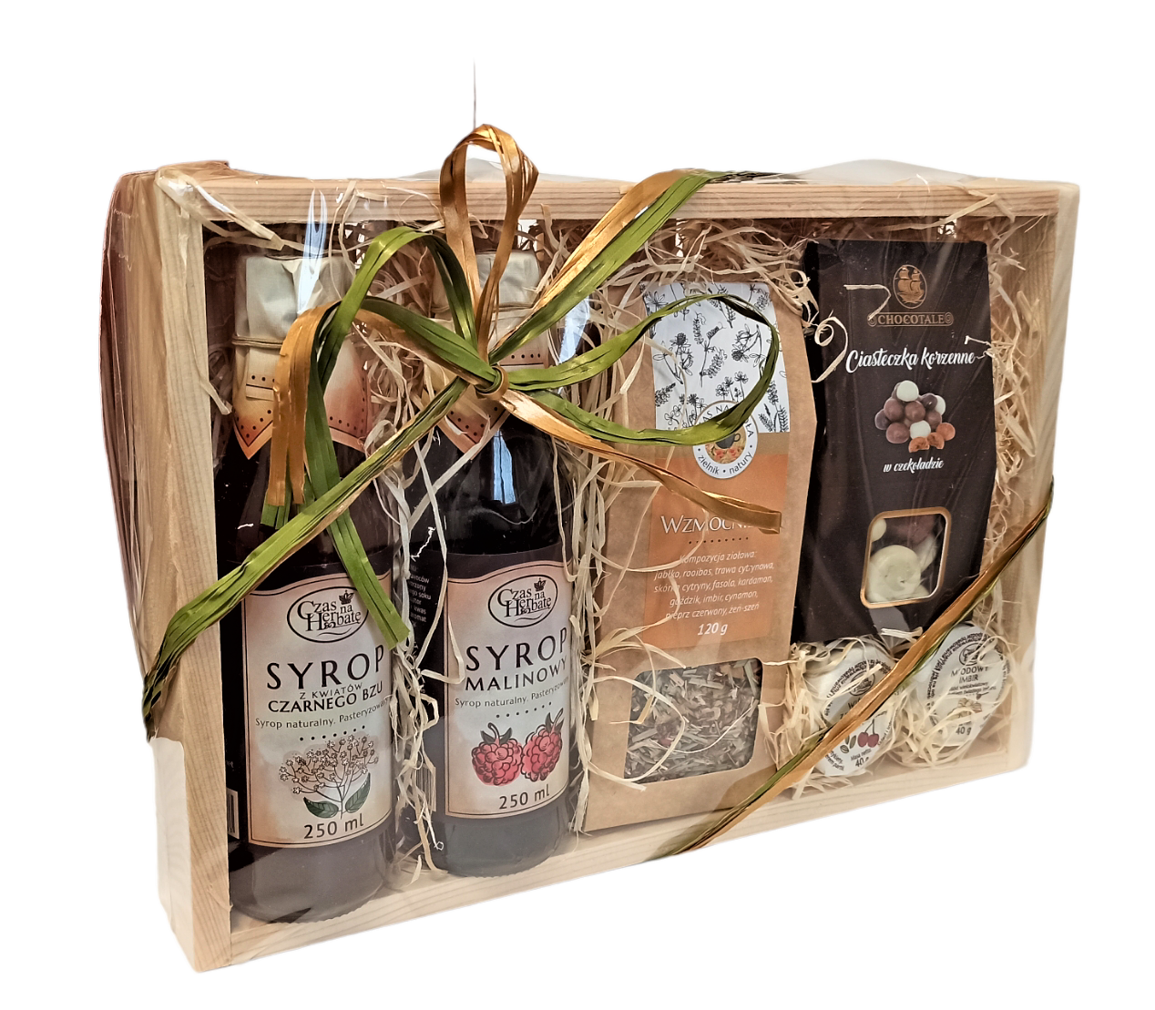 Widok z boku na drewnianą skrzynkę obwiązaną zieloną wstążką, w środku: syrop z kwiatów czarnego bzu w szklanej butelce, syrop malinowy w szklanej butelce, opakowanie herbaty, opakowanie ciasteczek korzennych w czekoladzie, dwa słoiczki