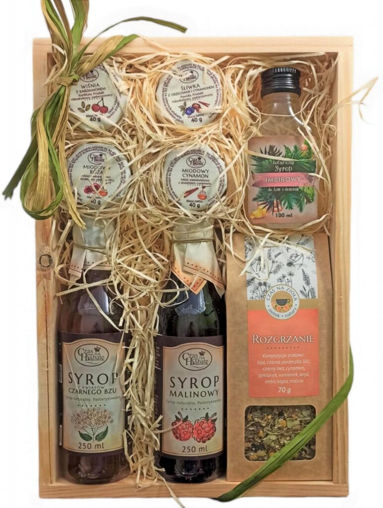 Drewniana skrzynka obwiązana zieloną wstążką, w środku: cztery słoiczki, butelka syropu imbirowego, syrop z kwiatów czarnego bzu, syrop malinowy oraz opakowanie herbaty