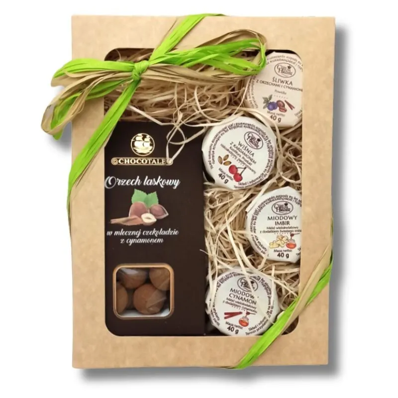 Beżowy kartonik z zieloną wstążką, w środku: opakowanie orzecha laskowego w czekoladzie, słoiczek miodowego cynamonu, słoiczek miodowego imbiru, słoiczek wiśni z kardamonem, słoiczek śliwki z orzechami i cynamonem