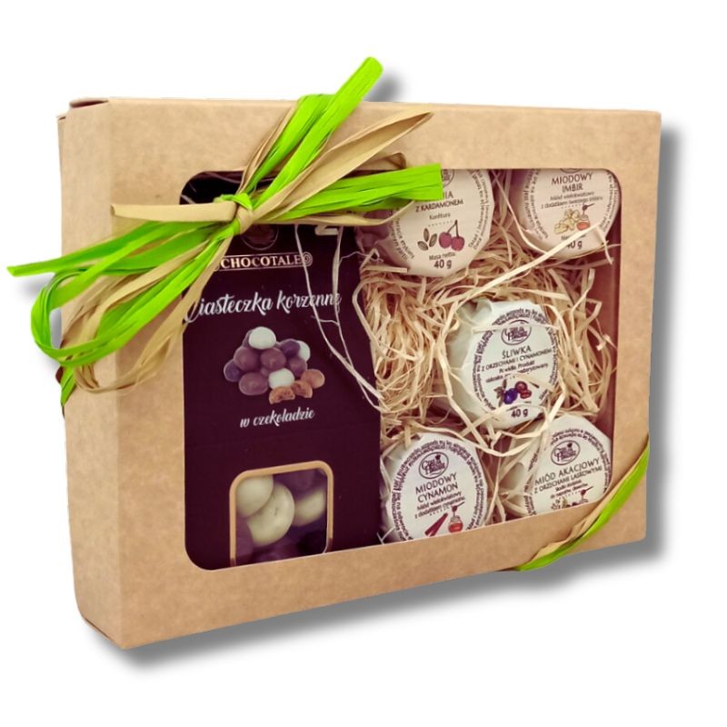 Widok z boku na beżowy kartonik obwiązany zieloną wstążką, w środku opakowanie ciasteczek korzennych oraz pięć słoiczków