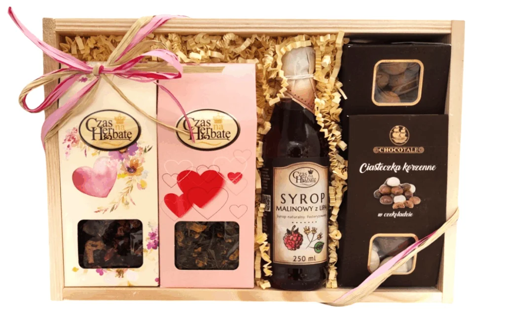 Drewniana skrzynka przewiązana różową wstążką, dwa opakowania herbaty ozdobione rysunkami serc, szklana butelka z syropem malinowym z lipą, opakowanie ciasteczek korzennych w czekoladzie i orzechów laskowych w czekoladzie z cynamonem