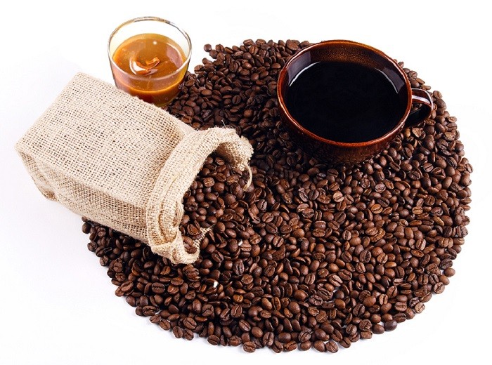 Ziarna kawy usypane w koło, a na nich brązowa filiżanka z kawą, jutowy woreczek i szklanka z karmelem