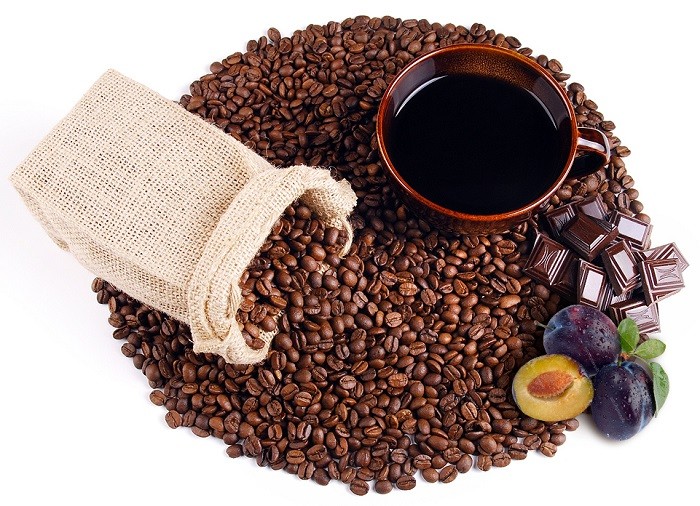 Ziarna kawy usypane w koło, a na nich brązowa filiżanka z kawą, jutowy woreczek, śliwki i czekolada