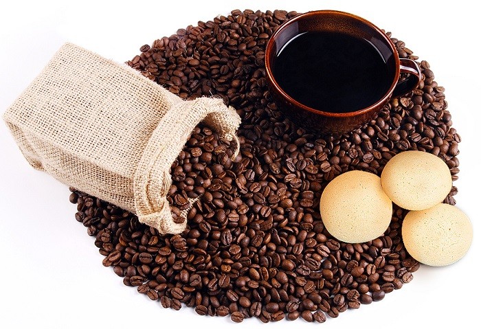 Ziarna kawy usypane w koło, a na nich brązowa filiżanka z kawą, jutowy woreczek i biszkopty