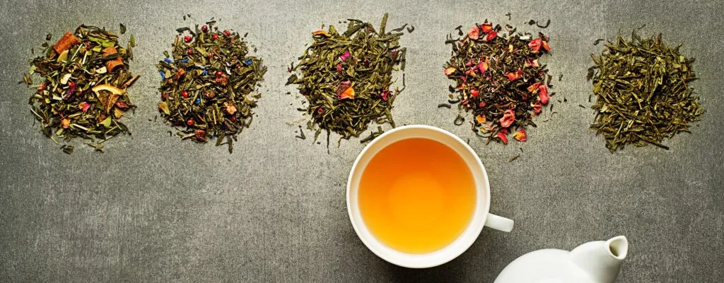 Pięć rodzajów sypanej herbaty ułożonych w rządek, filiżanka jasnego naparu i biały dzbanek na szarym tle.