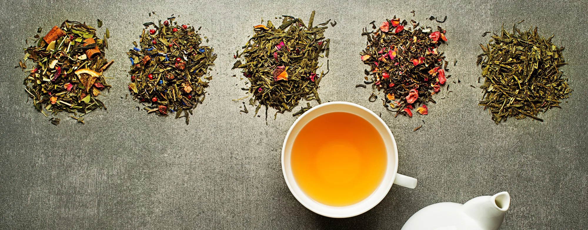 Pięć rodzajów sypanej herbaty ułożonych w rządek, filiżanka jasnego naparu i biały dzbanek na szarym tle.