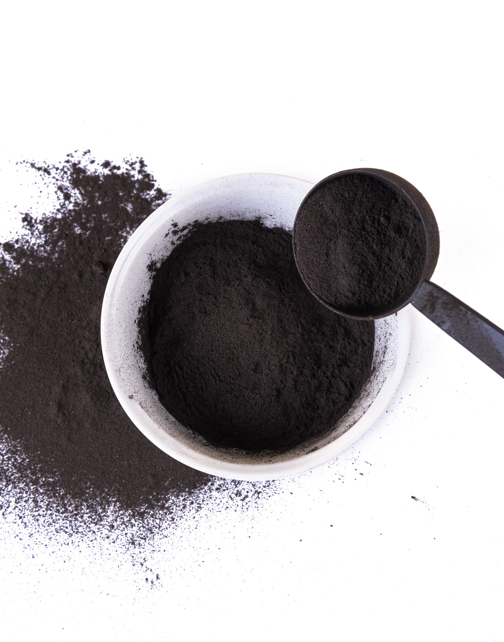 Biała miska z czarnym proszkiem, obok rozsypany proszek i czarna łyżka z porcją proszku.