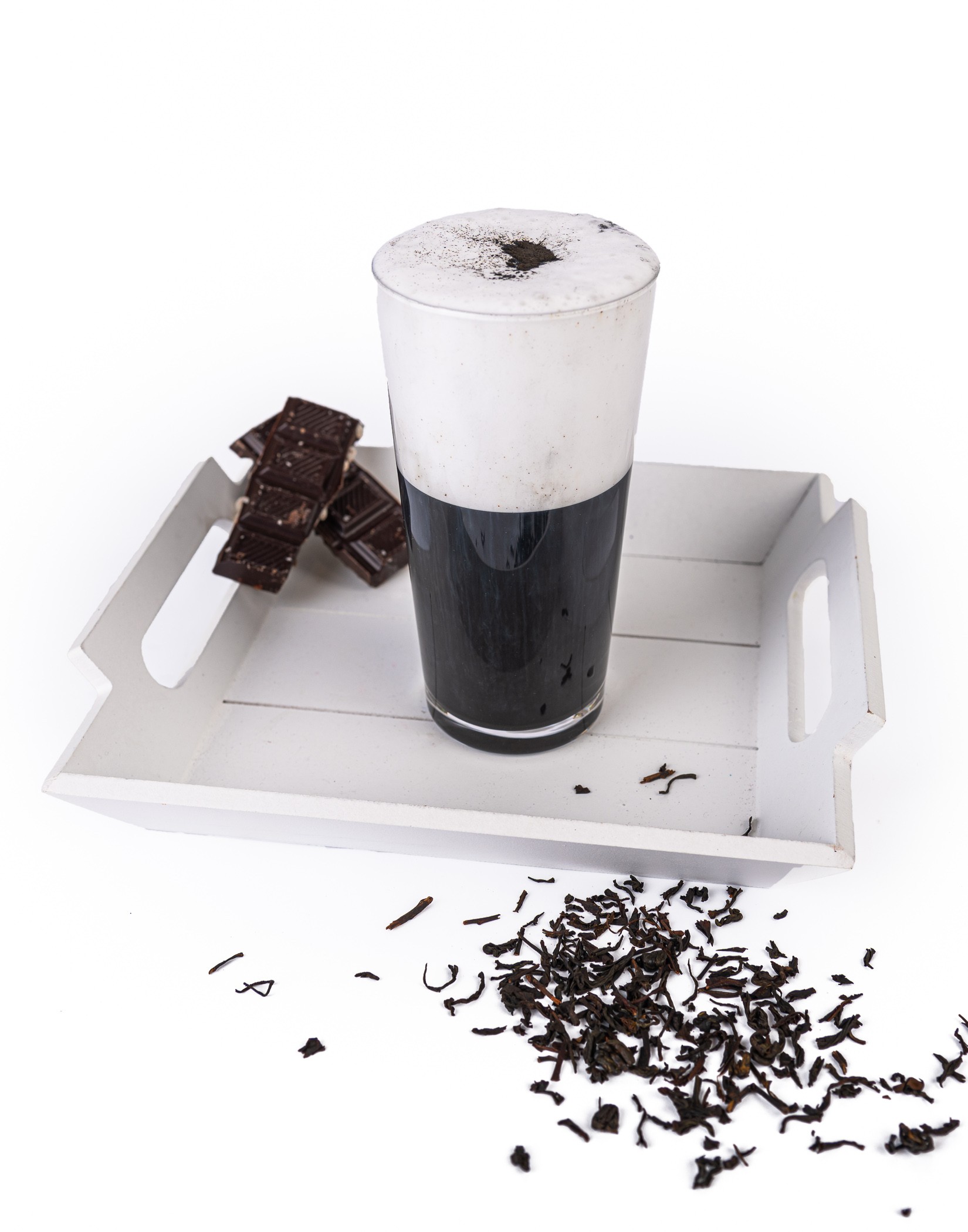 Szklanka czarnego napoju z pianką i posypką, na tacy obok kawałki gorzkiej czekolady i liście herbaty.